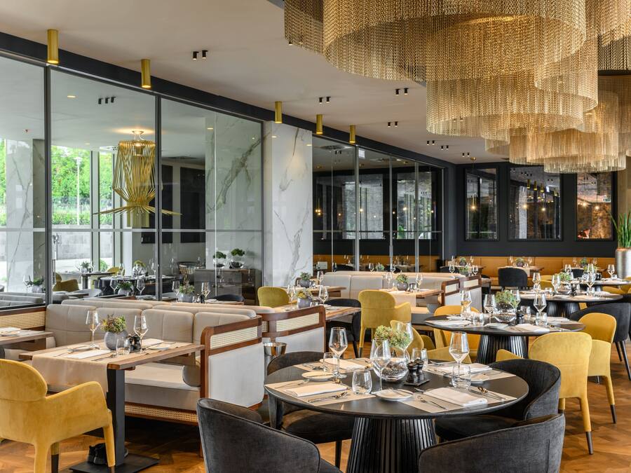 A modern restaurant interior with glass partitions, wooden furniture, and a central chandelier.