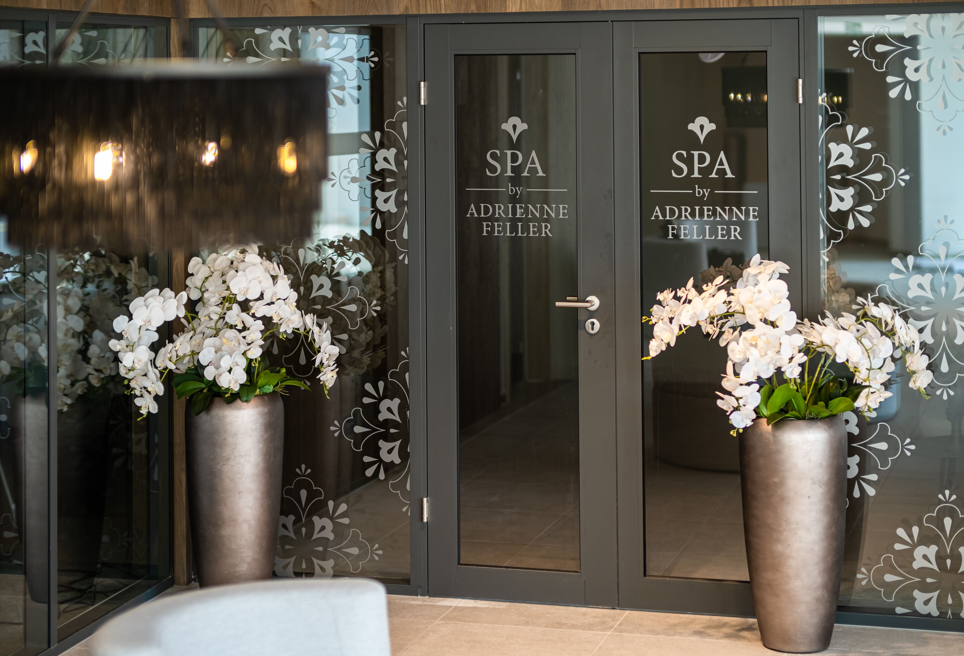 Entrada de un spa con dos plantas grandes en macetas y puertas de cristal con el rótulo 'SPA by Adrienne Feller'.