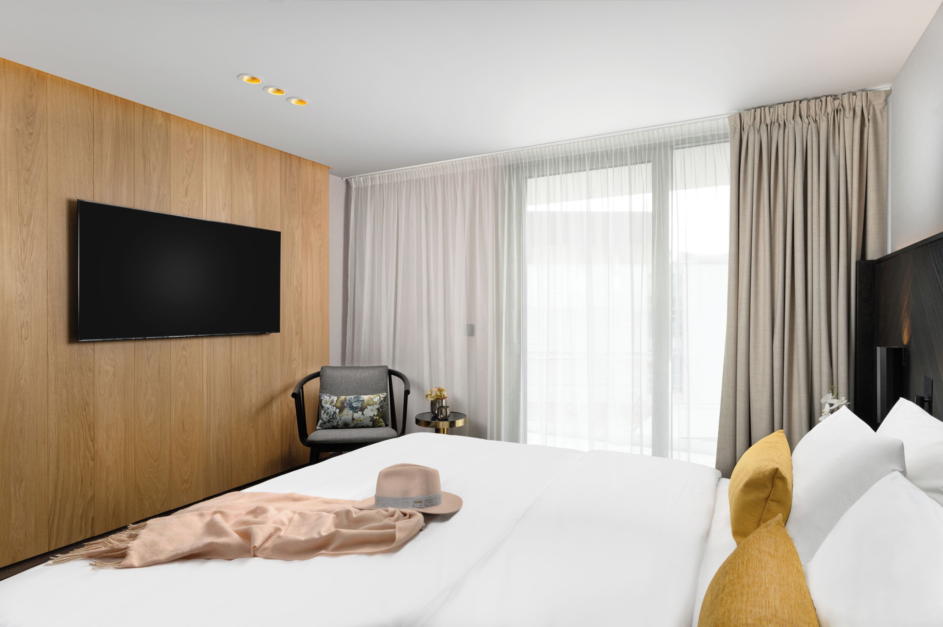 Une chambre d’hôtel moderne avec un grand lit, un téléviseur à écran plat fixé au mur, un fauteuil agrémenté d’un coussin à motifs floraux et une table de chevet sur laquelle est posé un vase de fleurs.