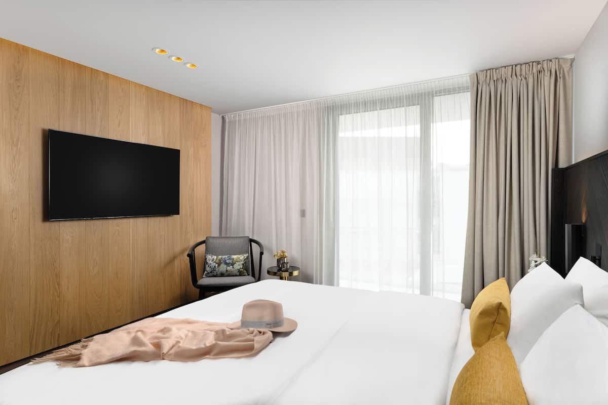 A modern hotel room with a large bed, a flat-screen TV mounted on the wall, a chair with a floral pillow, and a bedside table with a vase of flowers.