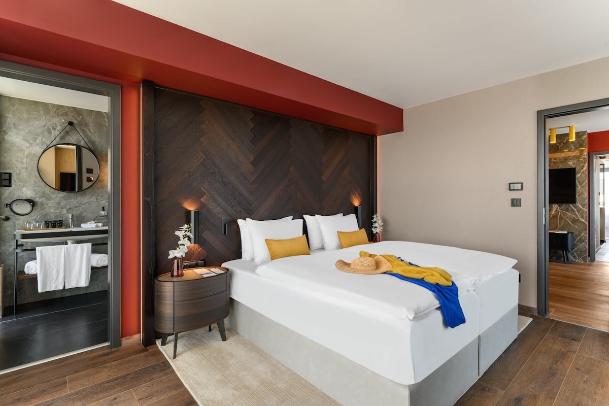 A modern hotel room with a large bed, a wooden herringbone headboard, a round mirror, a nightstand, and a bathroom with a marble countertop.
