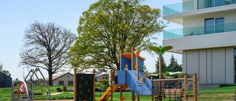 A playground with various play structures, a large tree, and a modern building in the background.