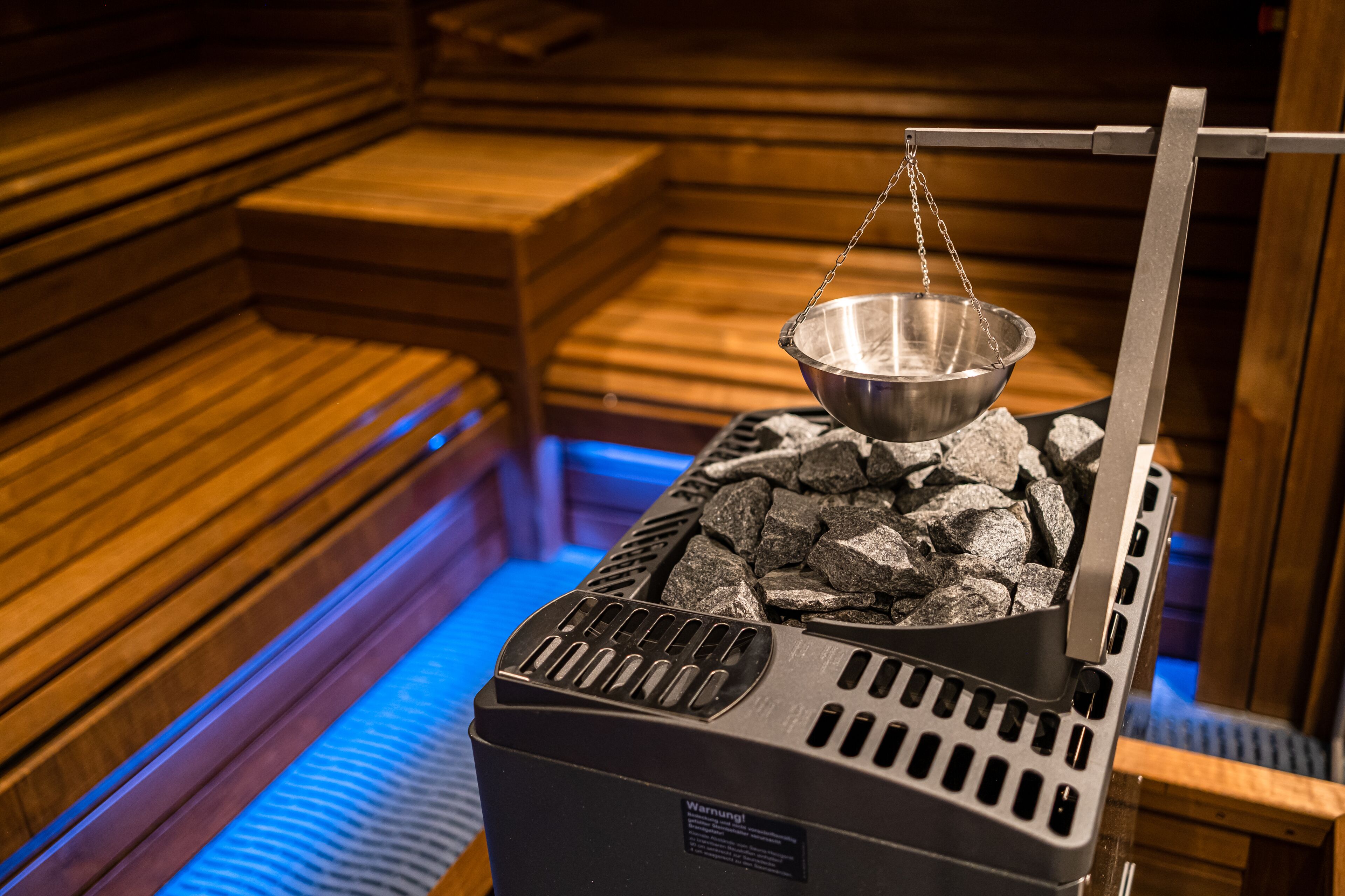A sauna with a metal bowl suspended over a bed of rocks, wooden benches, and a blue-lit floor.
