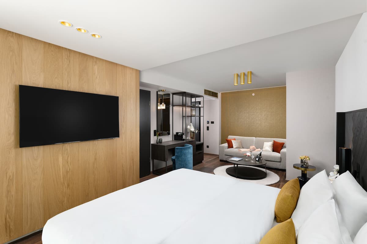 A modern hotel room with a large bed, a sofa, a desk, and a flat-screen TV mounted on the wall.