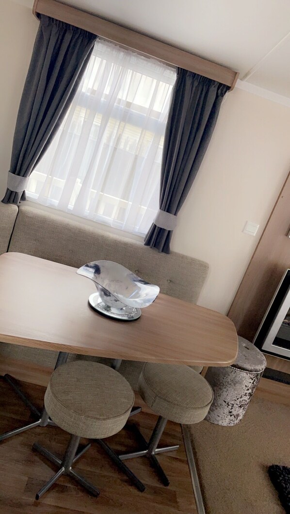 A dining area with a table, chairs, and a window with curtains. - Remarkable 3-bed Caravan, Direct Beach Access (Skegness)