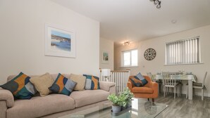 A living room with a beige sofa, orange armchair, glass coffee table, and a dining area with a white table and chairs. - Trewhiddle Retreat (St Austell)