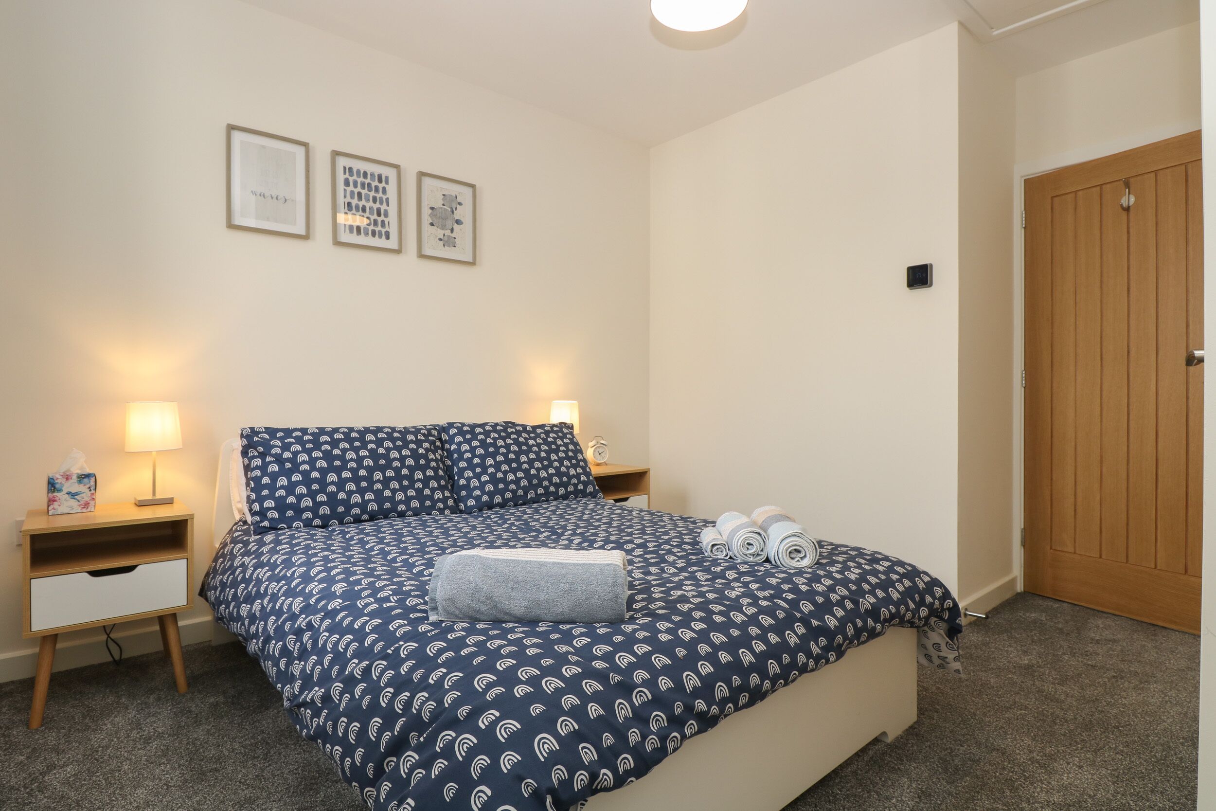 A neatly made bed with a patterned duvet, two bedside lamps, a small bedside table with a tissue box, and a wooden door.