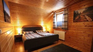 Tunnelmallinen, puinen mökkimainen makuuhuone, jossa on sänky, yöpöydät, ikkuna verhoilla ja patteri.