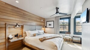 A modern bedroom with a wooden headboard, a bed with white bedding, a bedside table with a lamp, and a view of the outdoors through large windows. - The Carlin Washington | King bed, views (Breckenridge)