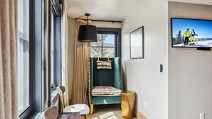 A cozy room with a large window, a green chair, a small table, and a mounted TV displaying a skiing scene. - The Carlin Washington | King bed, views (Breckenridge)
