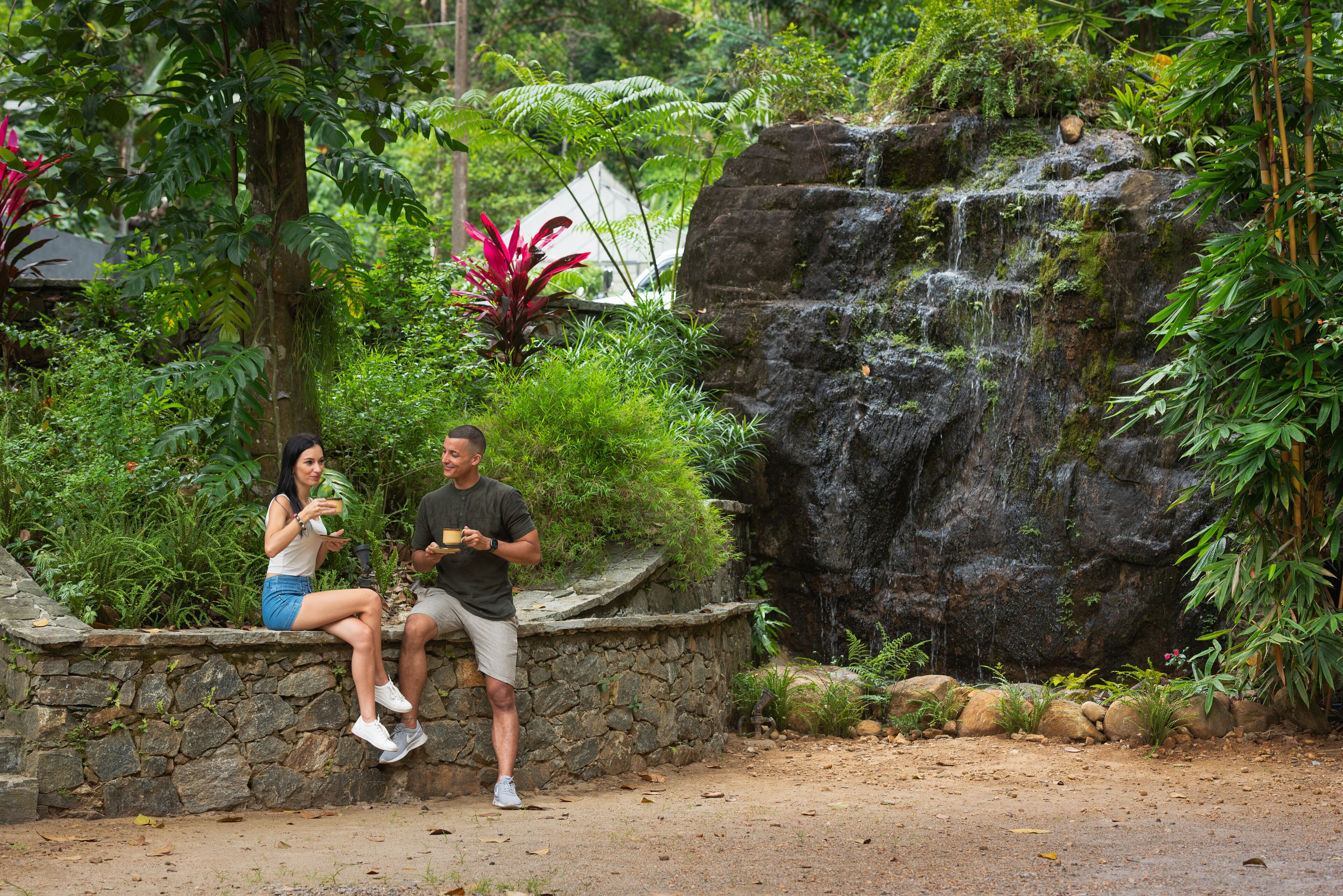 Una pareja sentada sobre un muro de piedra en un jardín exuberante, con una cascada y abundante vegetación.