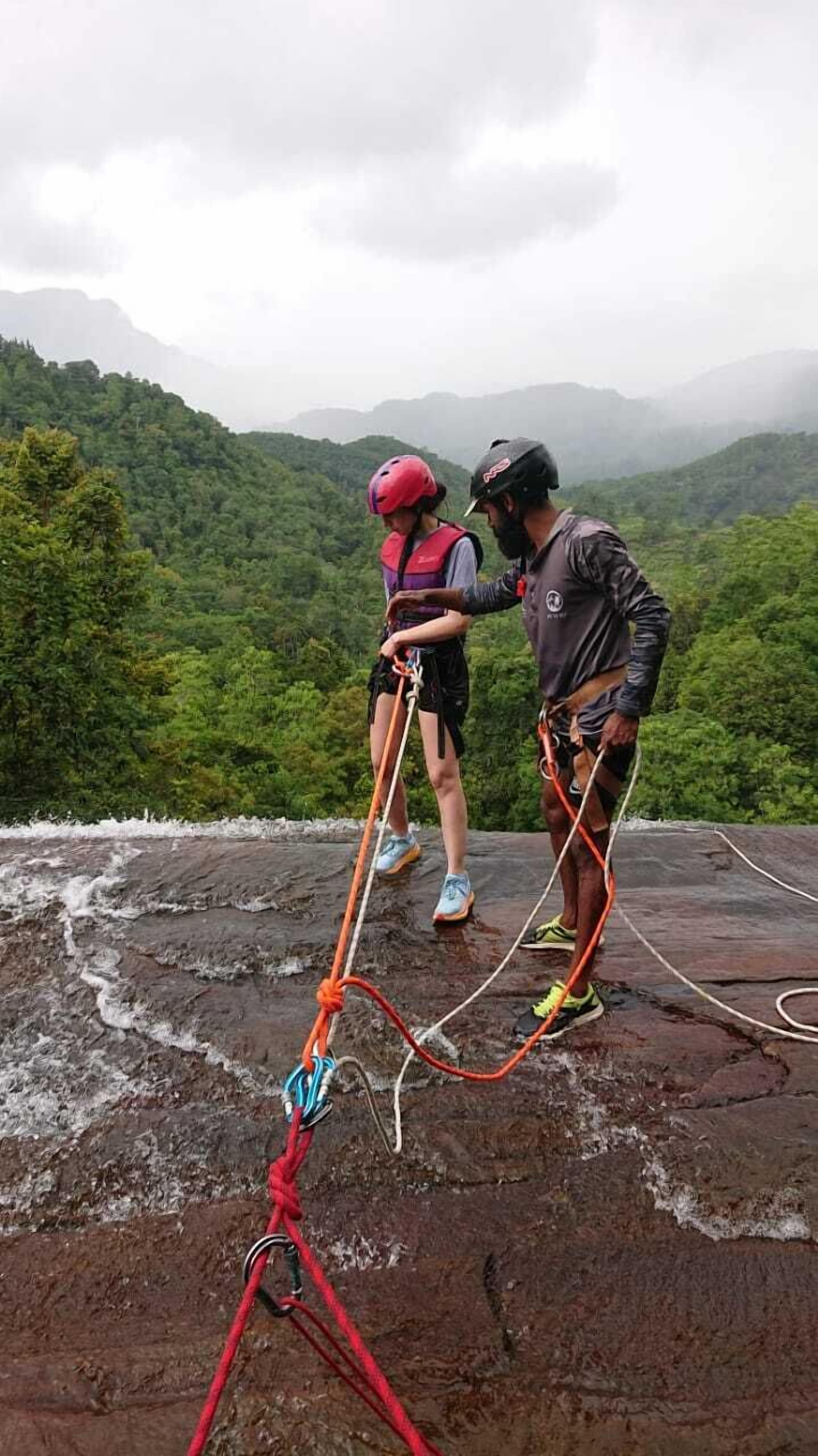 Zwei Personen auf einem felsigen Vorsprung mit Kletterseilen, umgeben von einer bewaldeten Berglandschaft.