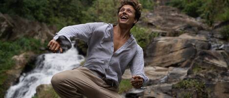 A person jumping in front of a waterfall with rocky terrain and greenery in the background.