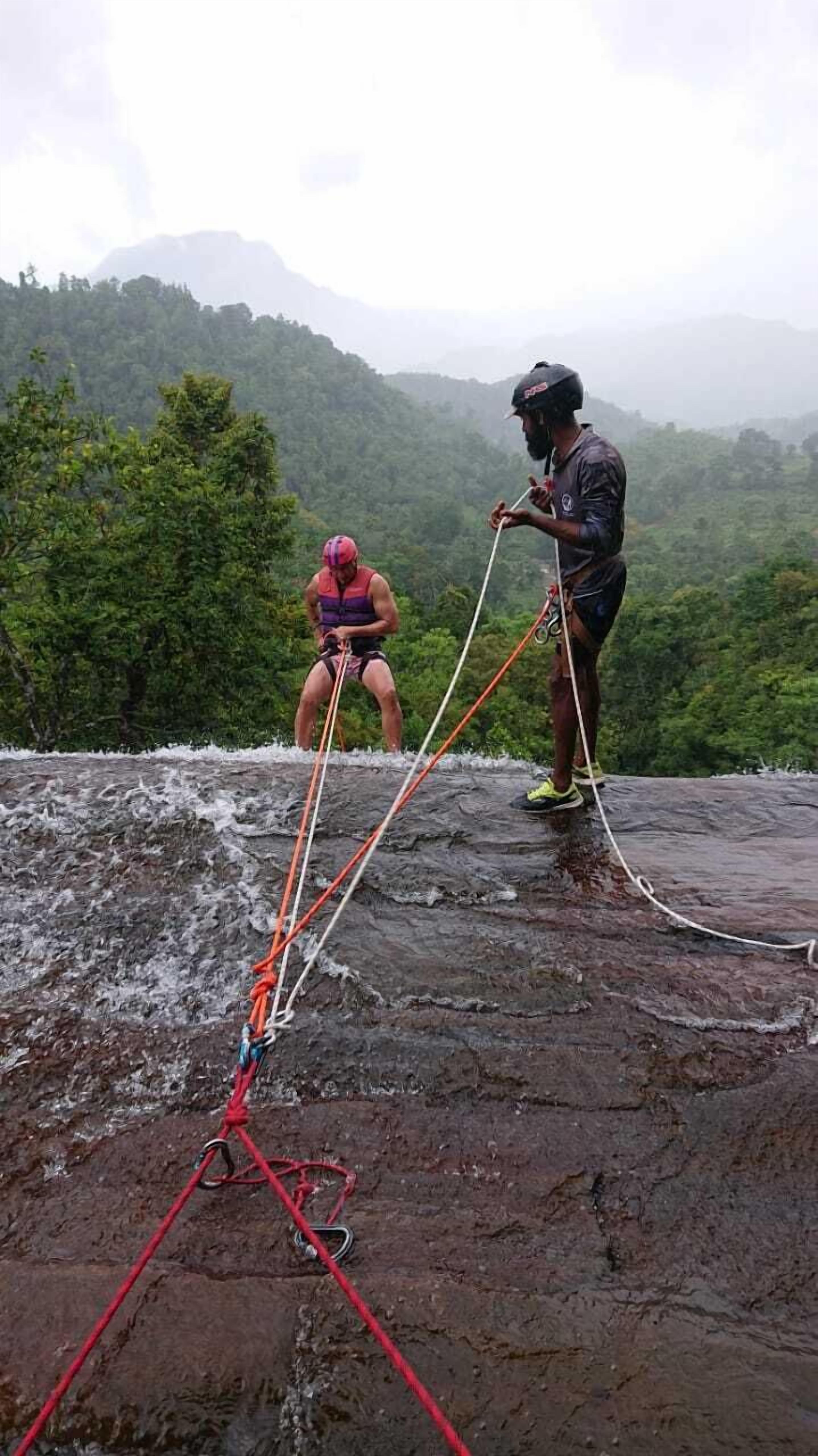 Two people rappelling on a wet rock surface with a forested mountain in the background.