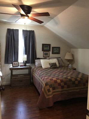 Quarto com cama, mesa de cabeceira, ventilador de teto e janela com cortinas.