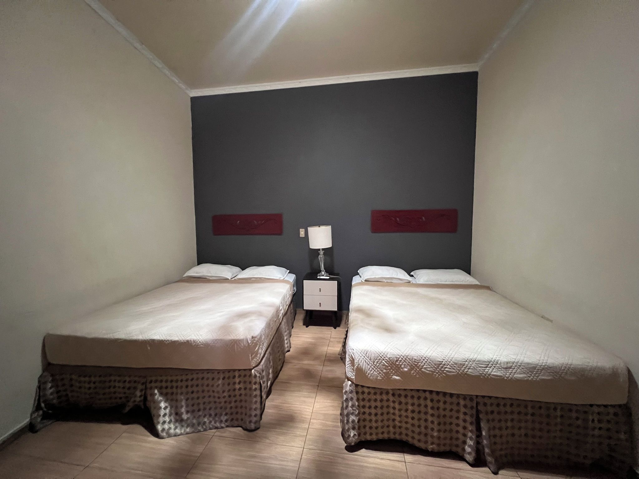 Una habitación con dos camas, una mesita de noche con lámpara y una pared de color oscuro.