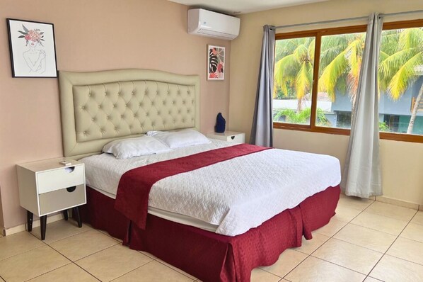 A hotel room with a bed, a nightstand, a wall-mounted air conditioner, and a window with curtains.
