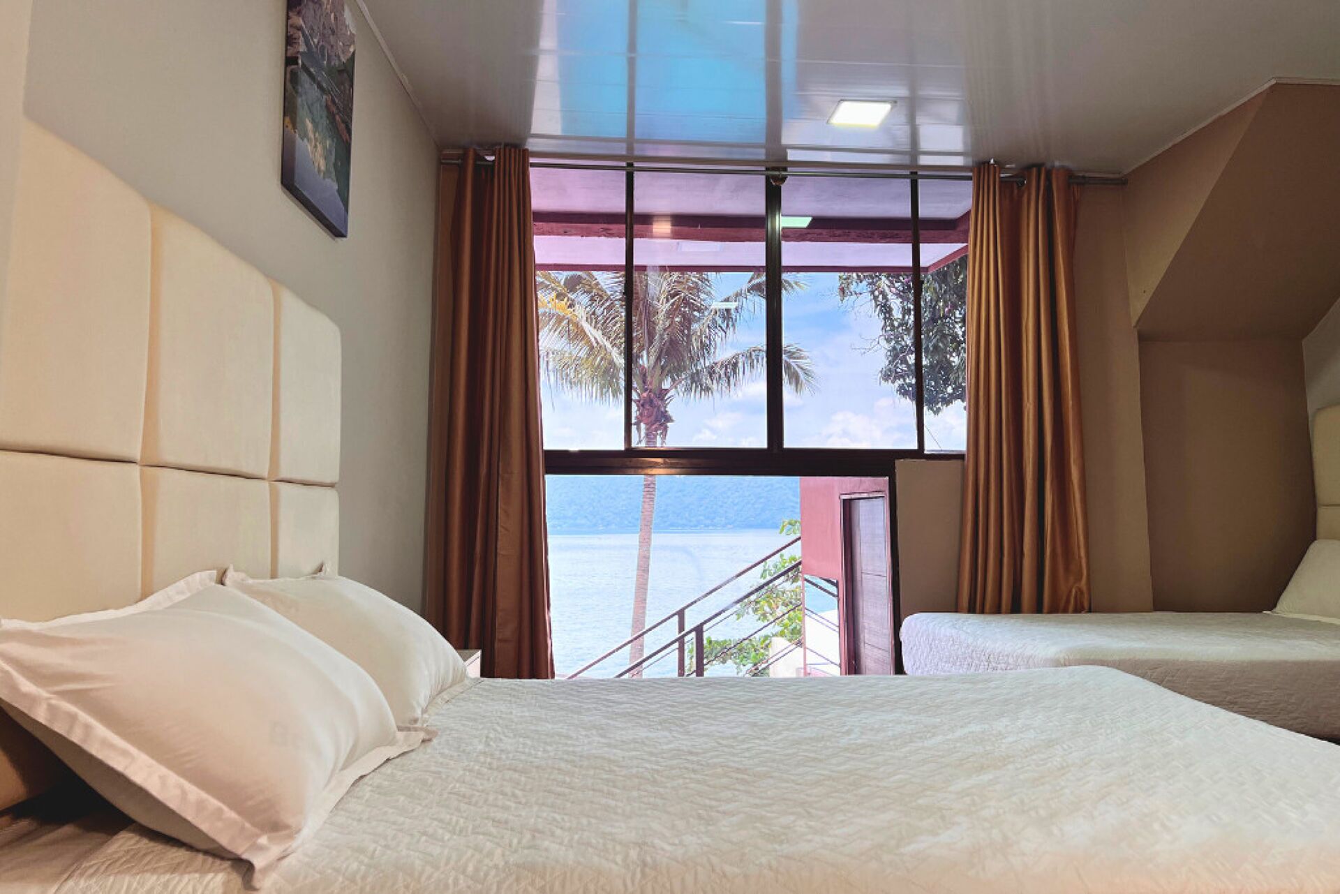 A hotel room with two beds, a large window with curtains, and a view of the sea.