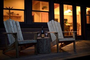 Two wooden Adirondack chairs on a wooden deck with a bottle and glasses on a stump.