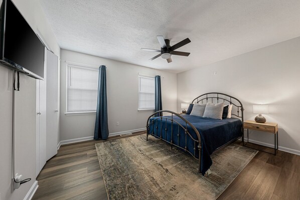 A bedroom with a bed, a ceiling fan, a nightstand with a lamp, and a TV mounted on the wall.