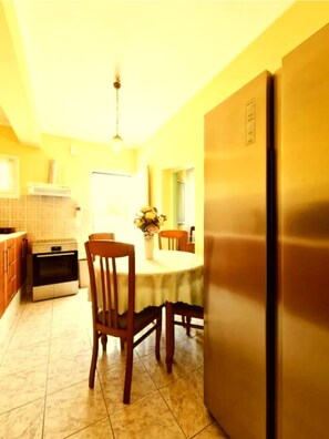 A kitchen with a refrigerator, oven, dining table, chairs, and a hanging light fixture.