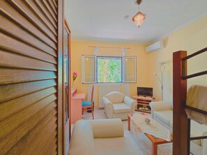 A room with a sofa, a table with a floral tablecloth, a television, and a window with a view of greenery. - Fully furnished tranquil apartment in Zakynthos (Lithakia)