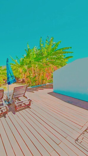A wooden deck with lounge chairs and a blue umbrella, surrounded by lush greenery.