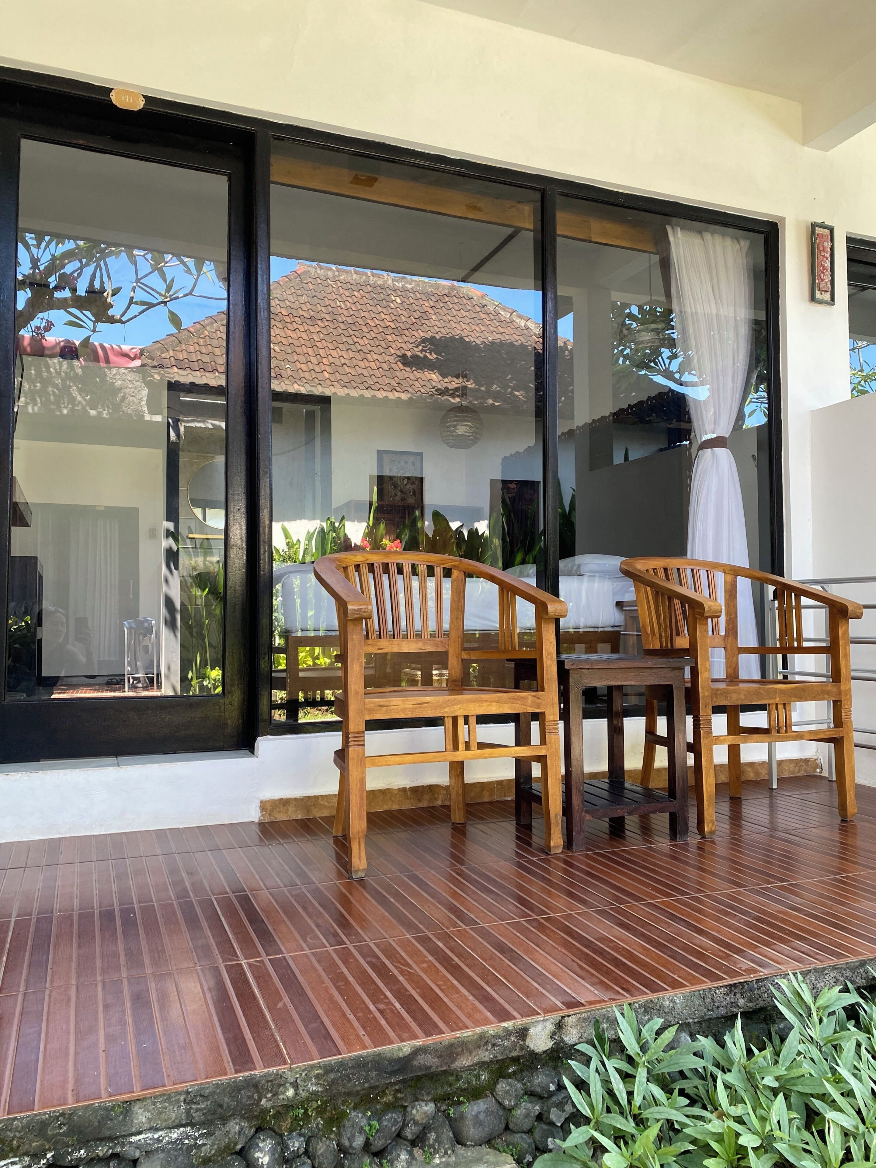 Una terraza de madera con dos sillas y una mesa, un vidrio corredero y vista a una casa con tejado de tejas.