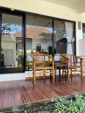 A wooden deck with two chairs and a table, a sliding glass door, and a view of a house with a tiled roof. - Alam Bali Homestay (Nusa Dua)