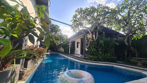 A swimming pool with a clear blue water, surrounded by lush greenery and a small building in the background.