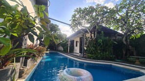 A swimming pool with a clear blue water, surrounded by lush greenery and a small building in the background. - Alam Bali Homestay (Nusa Dua)