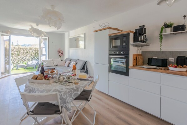 A modern kitchen with white cabinets, a built-in oven, and a coffee maker. There is a dining table with chairs and a small garden visible through the open door. - La Petite Poule Rousse - Maison avec piscine (Anduze)