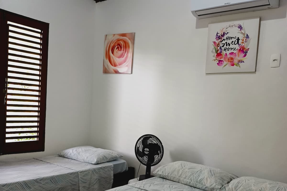 Une chambre avec deux lits, un ventilateur sur pied et une fenêtre avec des stores.