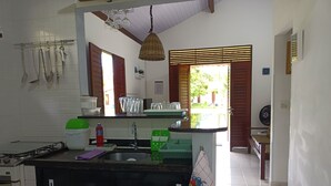 A kitchen with a gas stove, sink, and various utensils hanging on the wall. There is a window with wooden shutters, a hanging light fixture, and a view of the outside. - Recanto Feliz (Tamandaré)