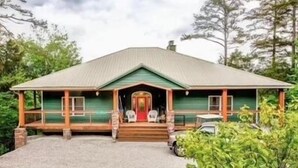 Een huis met twee verdiepingen, met een groene gevel, een veranda met twee stoelen en een auto ervoor geparkeerd.