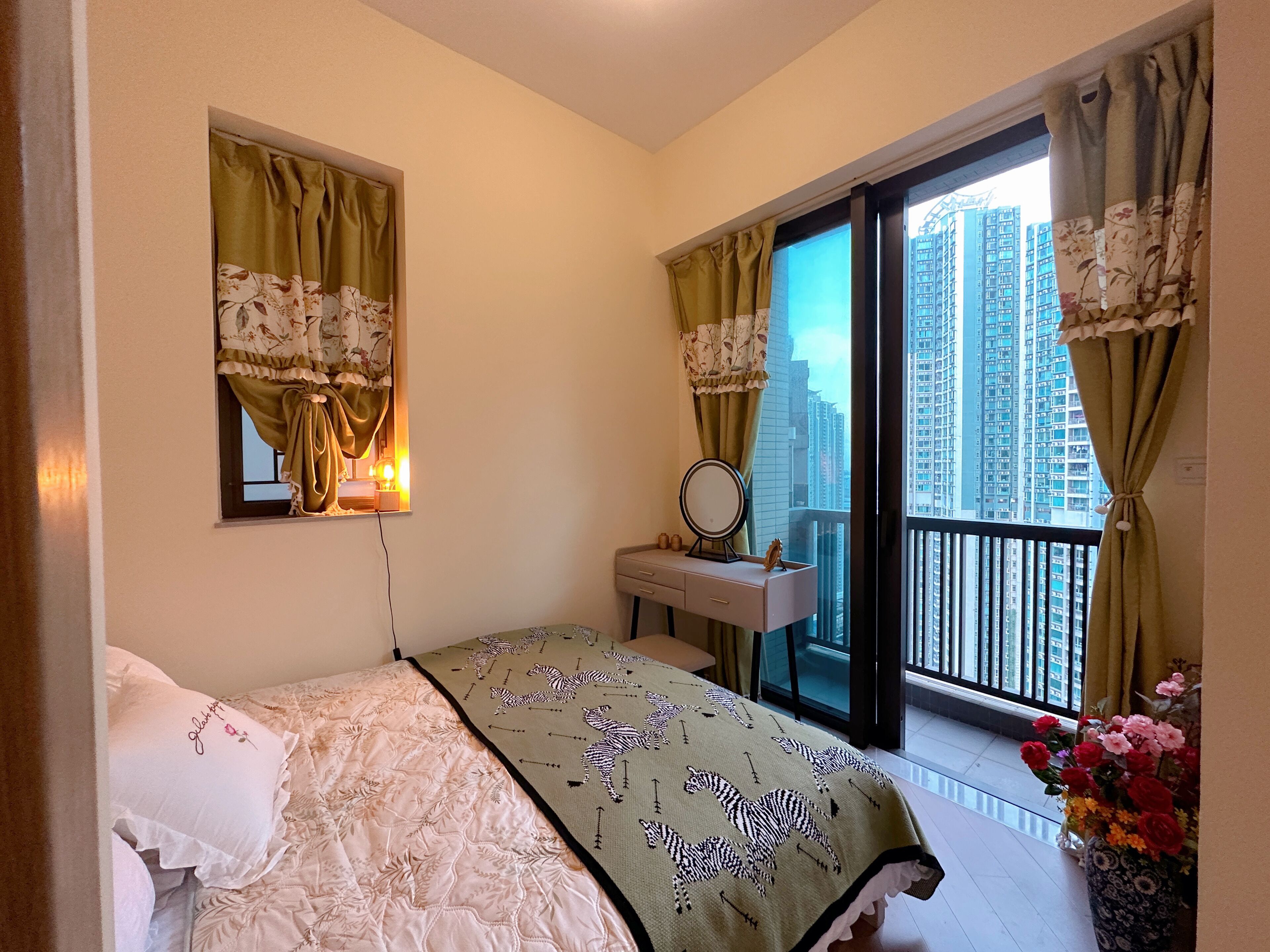 Un dormitorio con una cama, una tocador con espejo, una ventana con cortinas y un balcón con vistas a edificios altos.