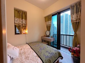 Un dormitorio con una cama, una tocador con espejo, una ventana con cortinas y un balcón con vistas a edificios altos.
