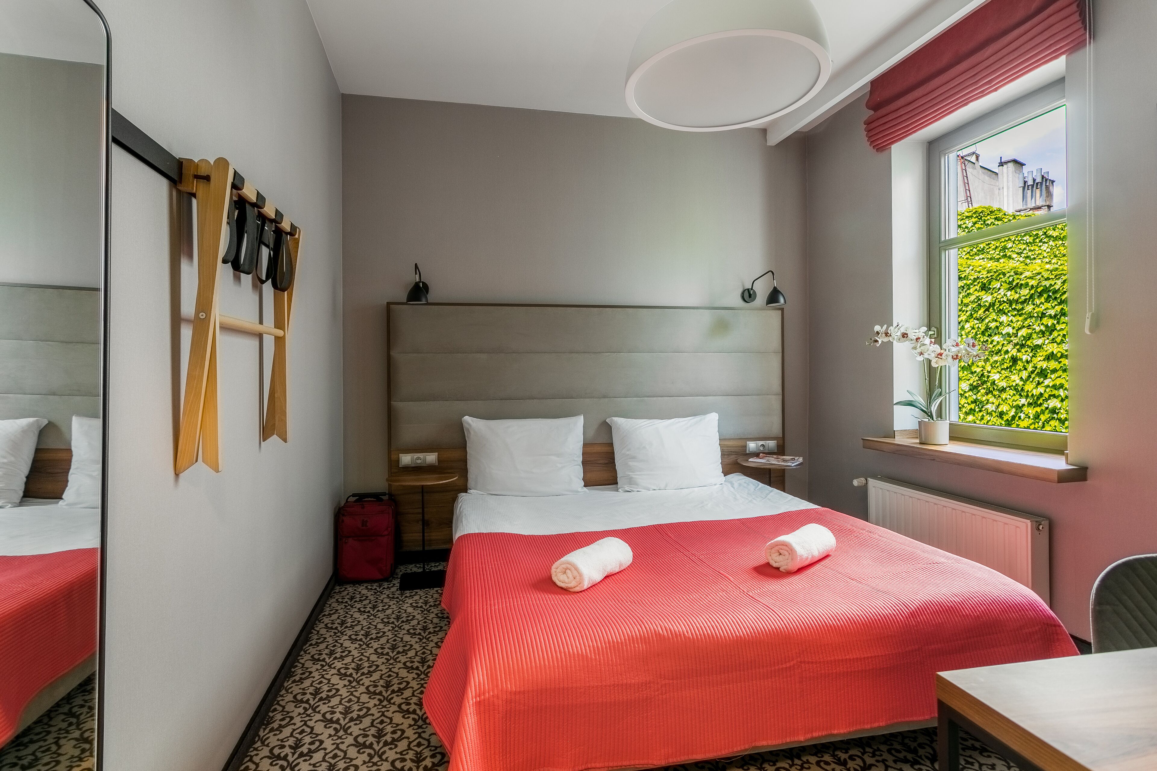A hotel room with a bed, a small table, a window with a view of greenery, and a mirror on the wall.