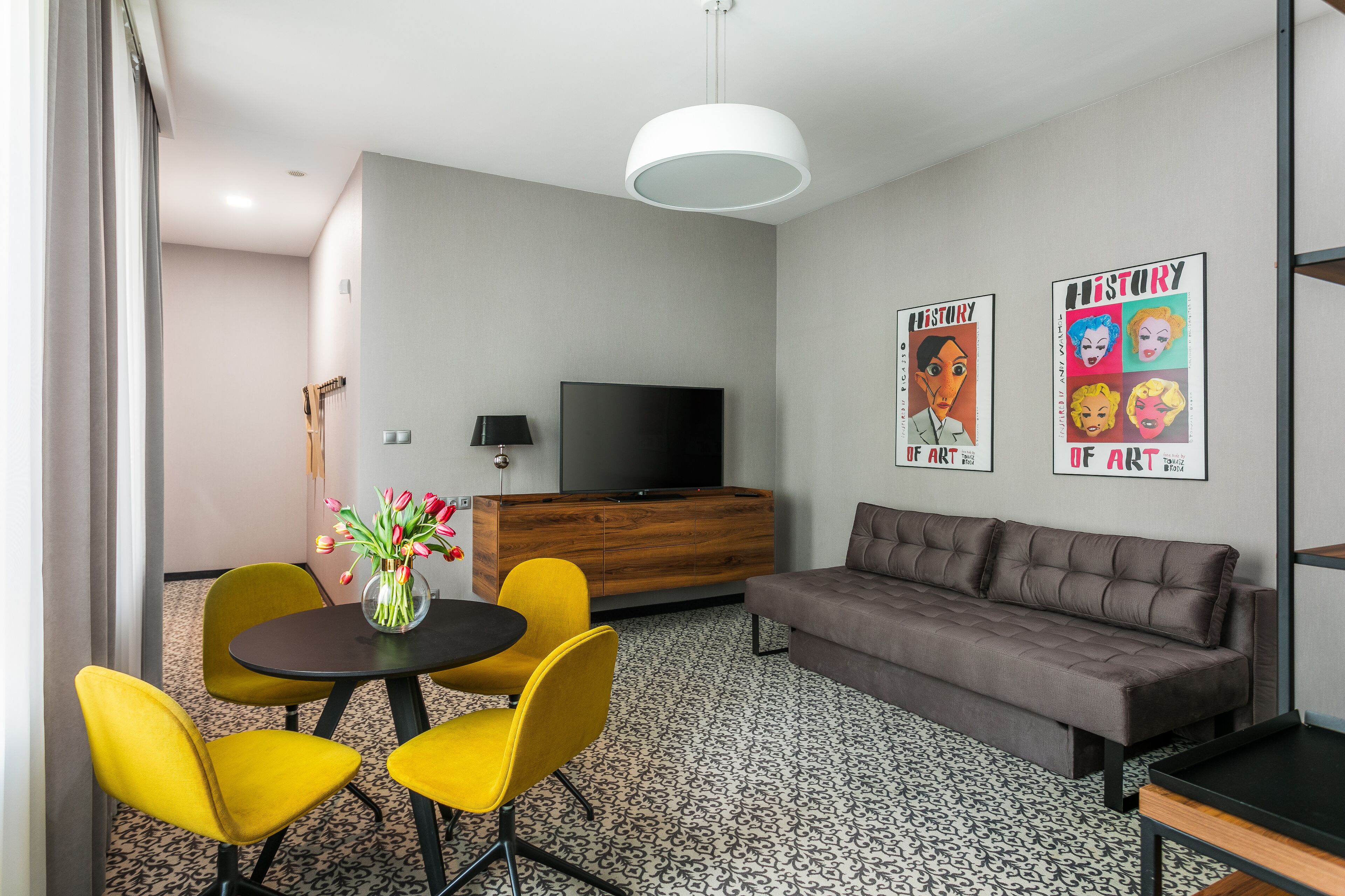 Una sala de estar moderna con un sofá gris, sillas amarillas, un mueble de madera, un televisor de pantalla plana y una lámpara colgante.