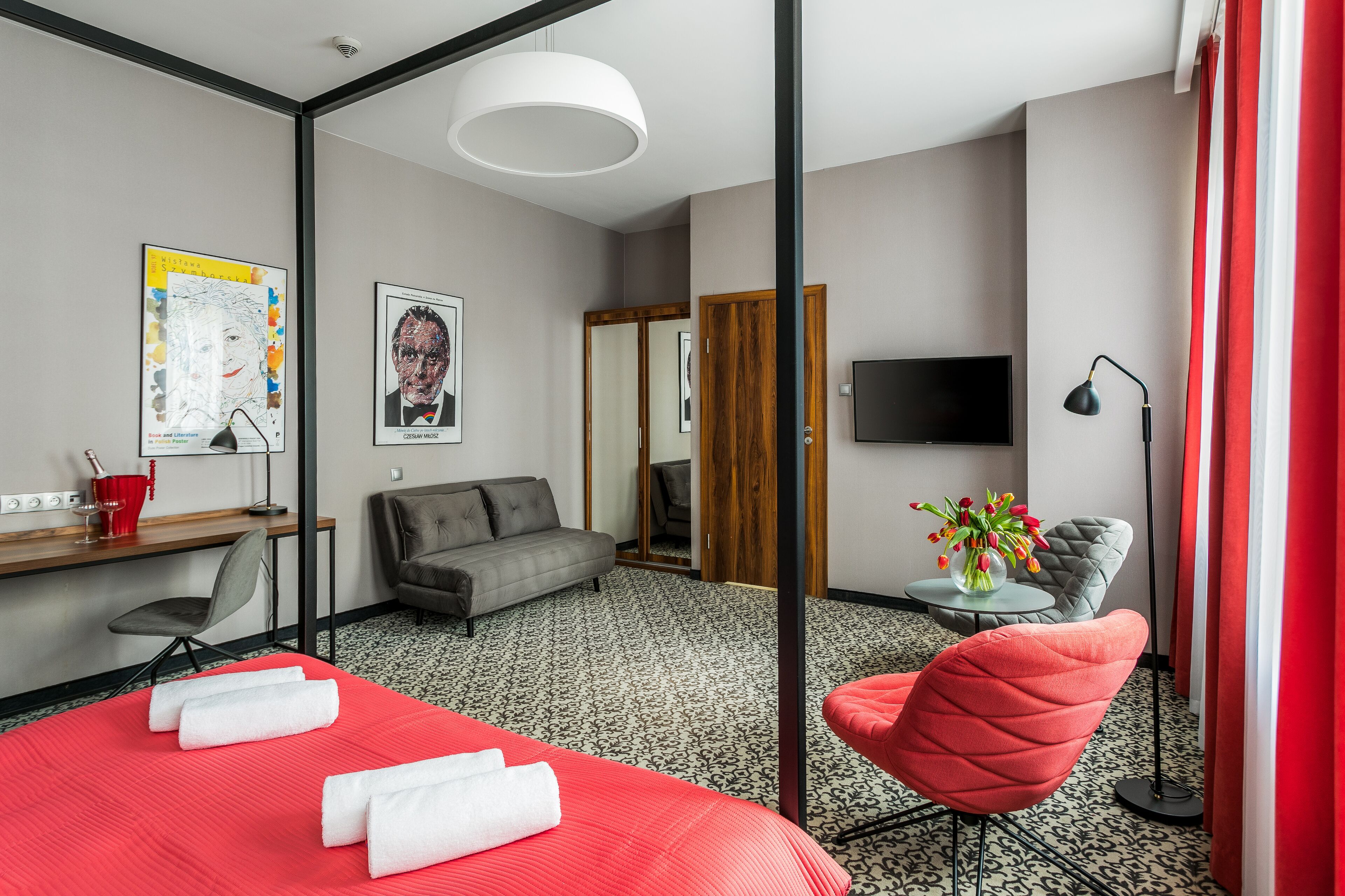 A hotel room with a red bed, grey sofa, a small table with a vase of flowers, a desk with a chair, a TV, and a mirror.