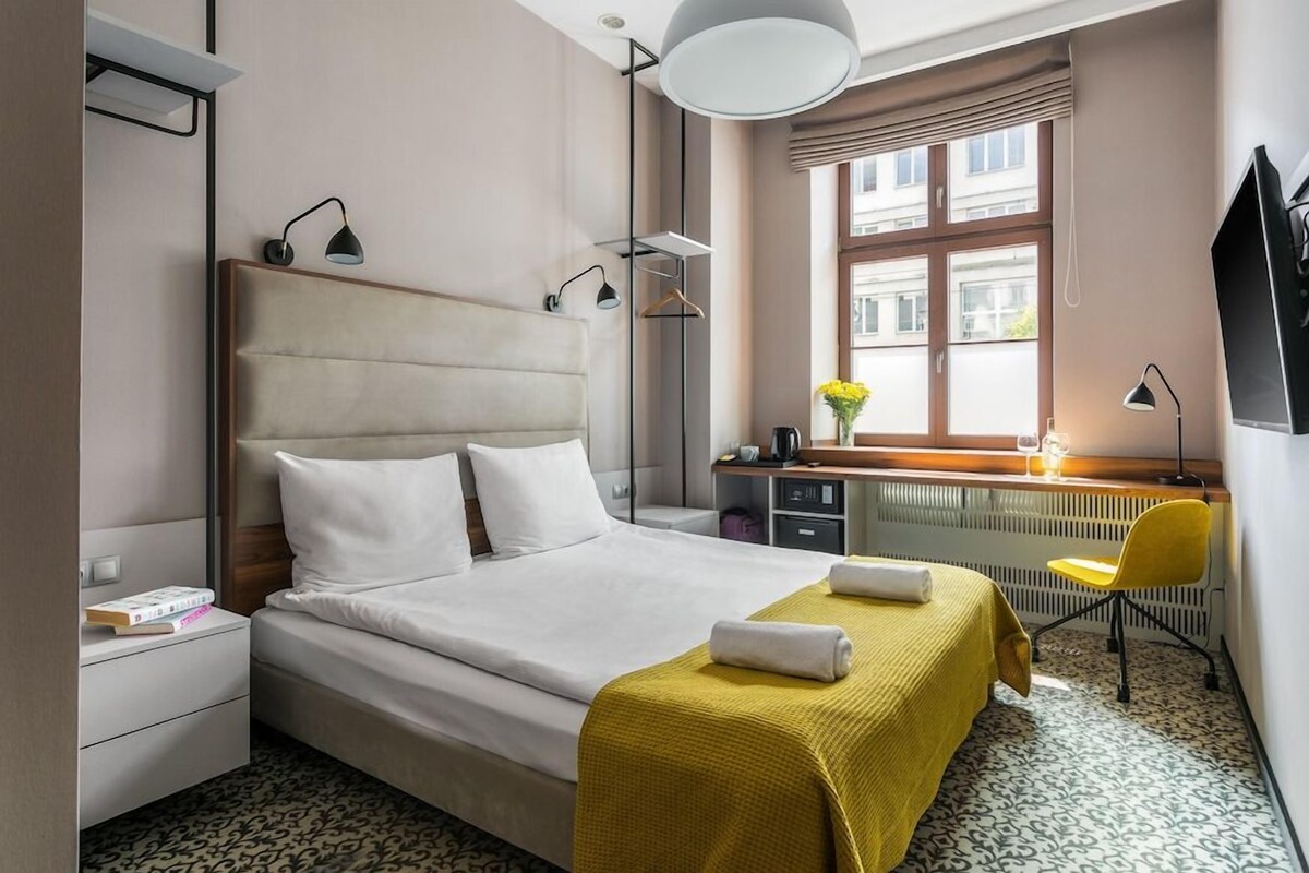 A hotel room with a bed, bedside tables, a desk with a chair, a radiator, and a window with a view of buildings.