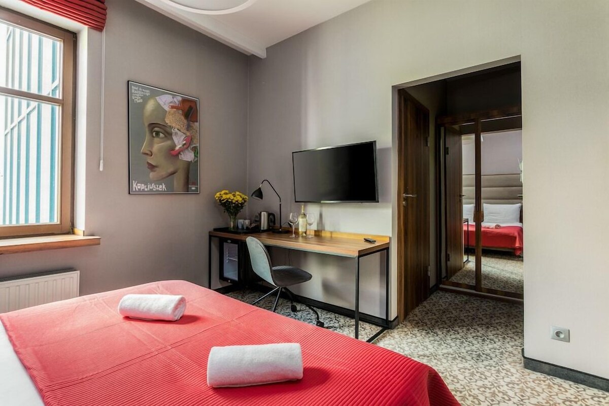 A hotel room with a bed, a desk with a computer, a TV, and a door leading to another room.