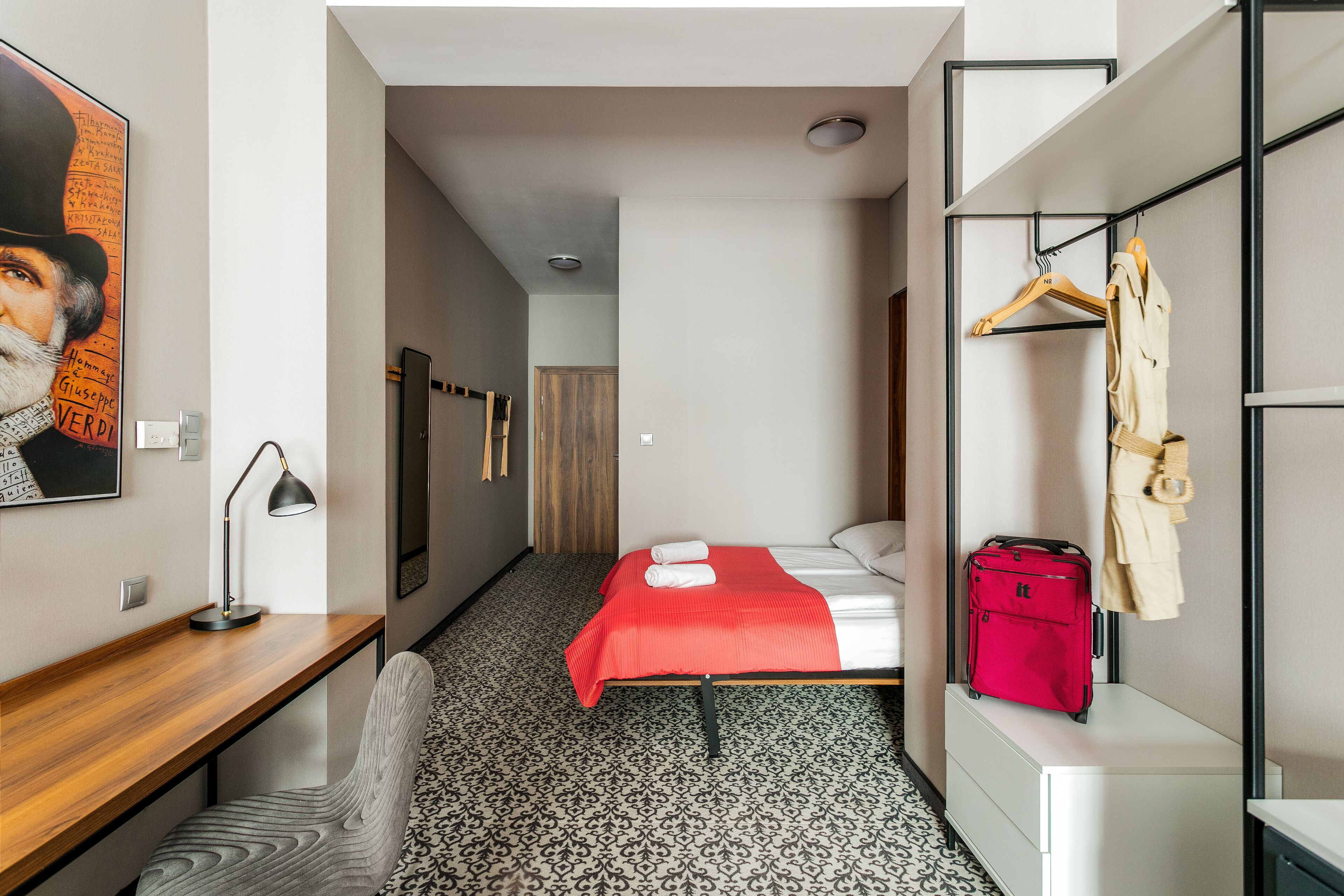Habitación de hotel con cama, escritorio, silla, armario y maleta roja.