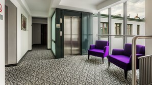 A hallway with patterned carpet, purple seating, an elevator, and large windows with a view of buildings. - No.23 Premium Aparthotel (Kraków)