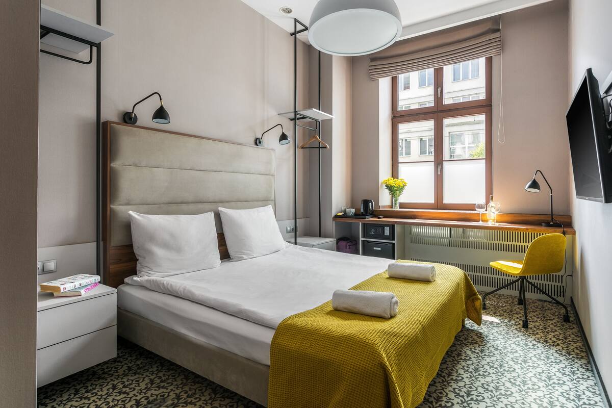 Habitación de hotel con cama, mesitas de noche, escritorio, silla, radiador y ventana con vistas a edificios.