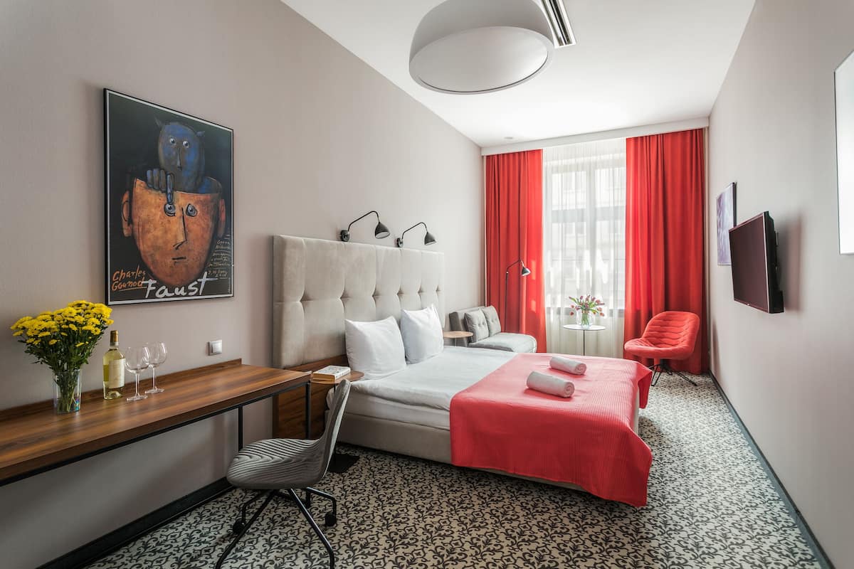 Una habitación de hotel moderna con una cama grande, un escritorio con un florero, una silla, una mesita con una lámpara y un cuadro en la pared.