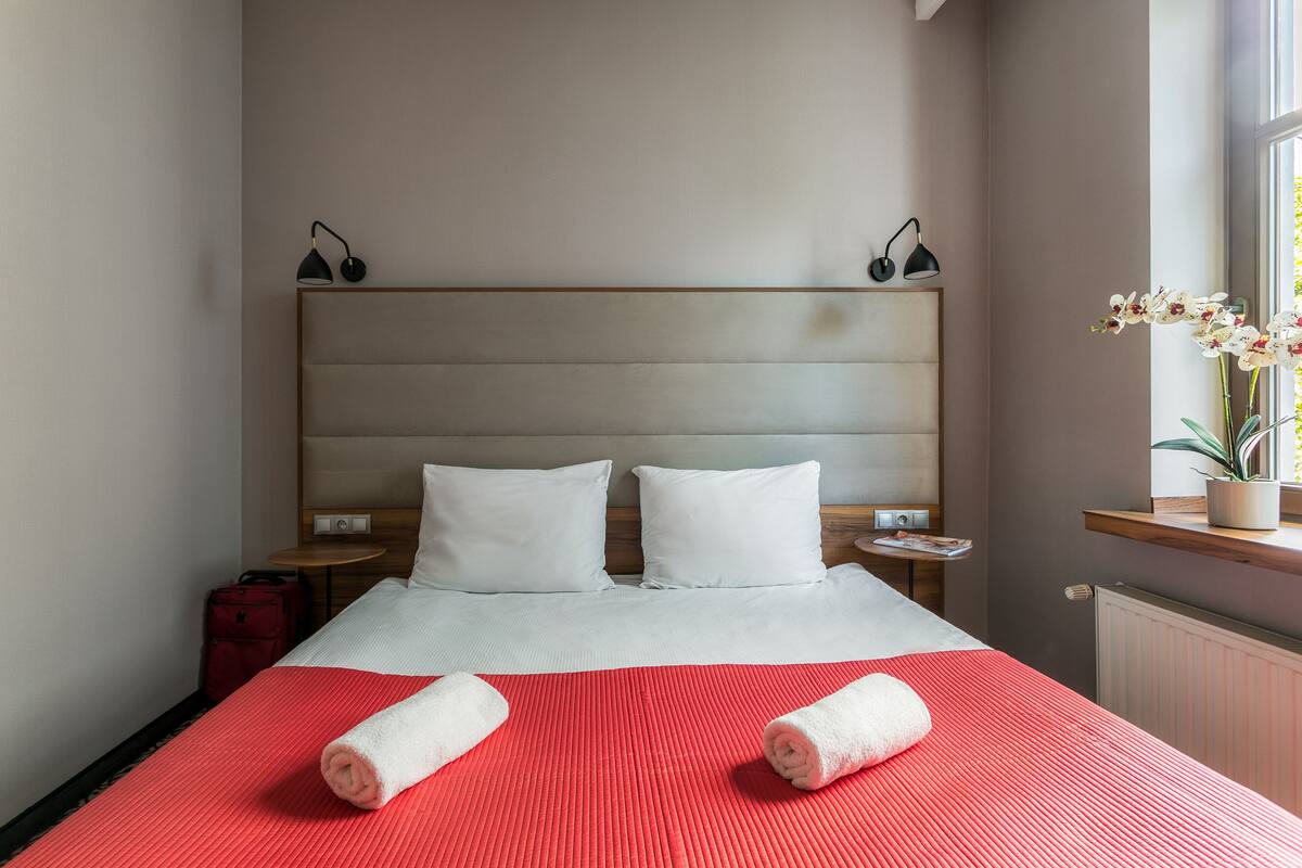 Habitación de hotel con una cama grande, dos almohadas blancas, una colcha roja, dos toallas enrolladas, una mesita de noche con lámpara, una maleta roja y una ventana con una planta de orquídea.