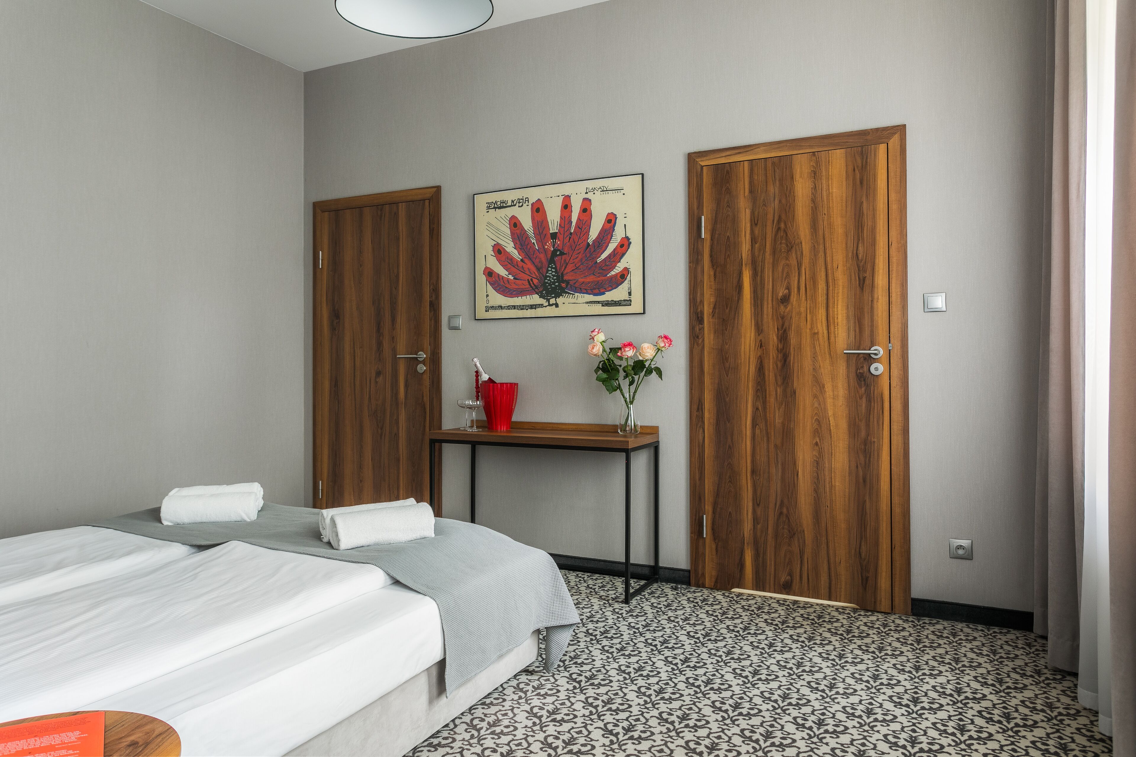 Une chambre d’hôtel avec deux lits, une porte en bois, une œuvre d’art encadrée, une petite table avec un vase et une tasse rouge, et un tapis à motifs.