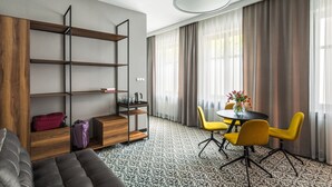 A modern hotel room with a sofa, a wooden bookshelf, a dining table with yellow chairs, and a large window with curtains. - No.23 Premium Aparthotel (Kraków)