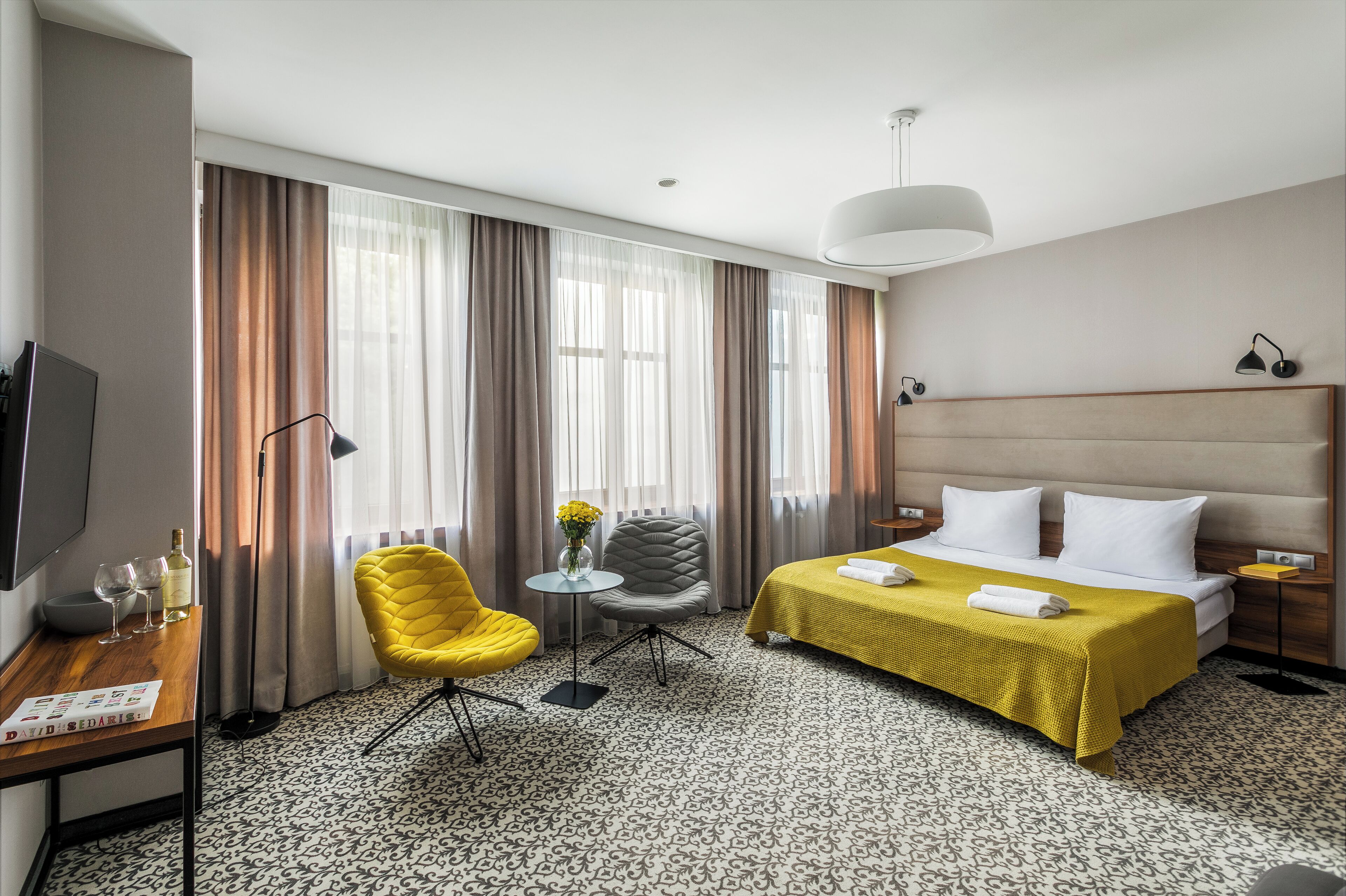 A hotel room with a bed, a desk, a chair, a small table with a vase, and curtains.